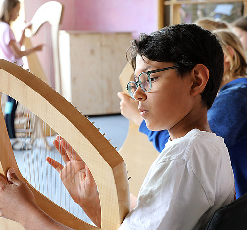 Ein Junge macht Musik mit einer Harfe und im Hintergrund sitzen weitere Schüler mit Instrumenten
